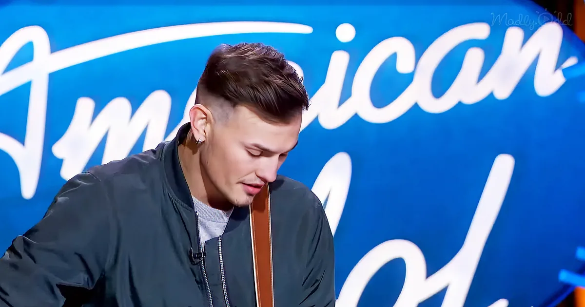 A man in a dark jacket holds a guitar, looking down, with an "American Idol" backdrop behind him.