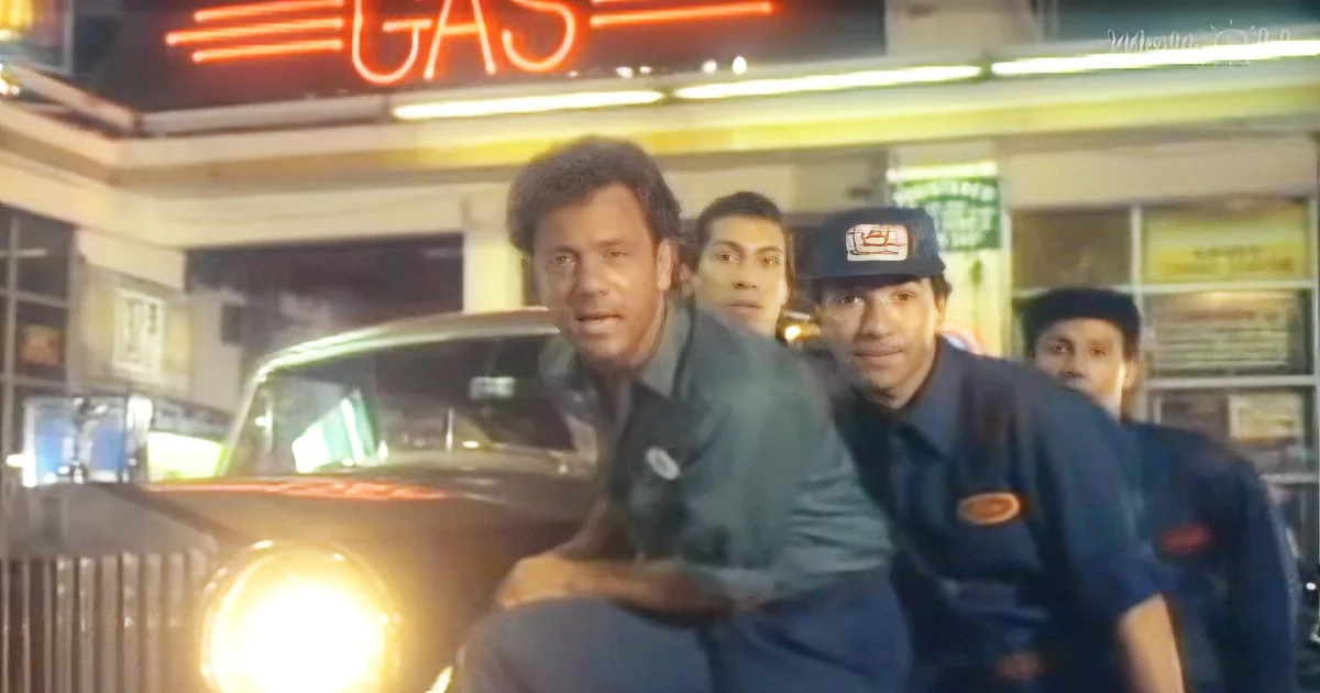 A man in a gas station uniform leans forward, looking tense, with a car's bright headlights in the foreground.