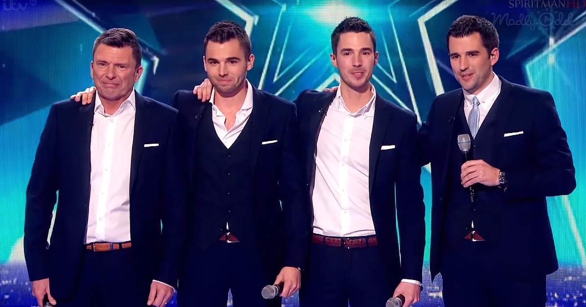 Four men in suits stand on stage, one holding a microphone, with a blue geometric background.