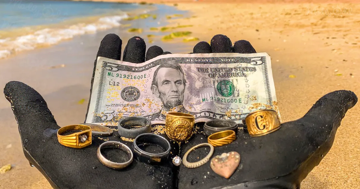A gloved hand holds a $5 bill and various rings, including a gold one with a "C," on a sandy beach.