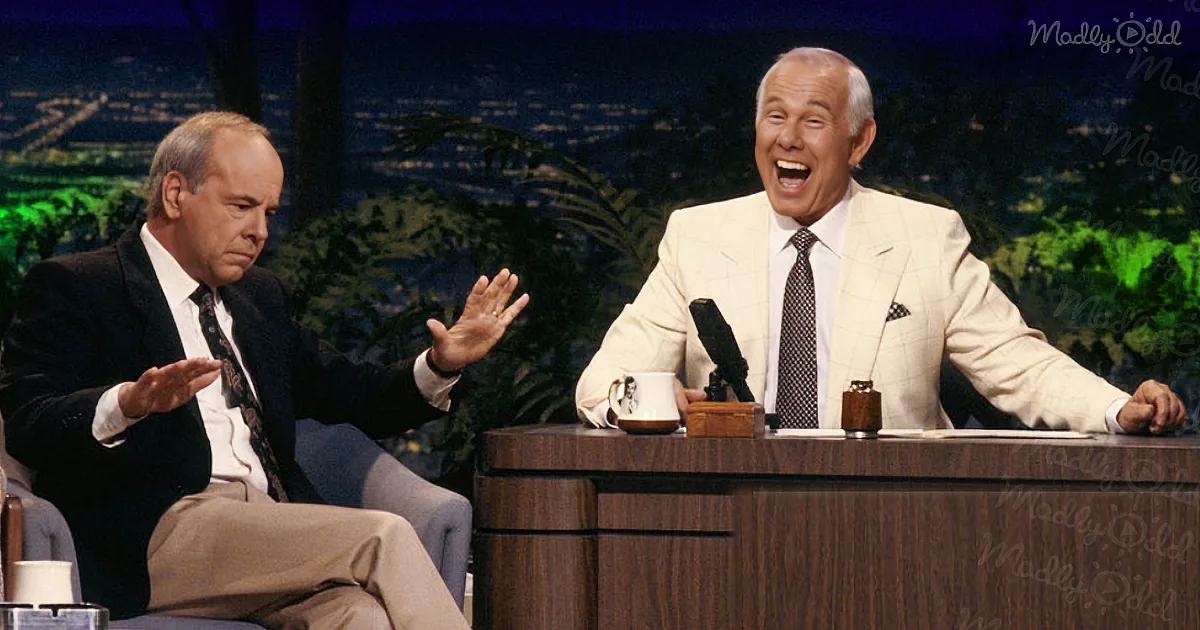 A man in a suit gestures animatedly while seated next to another man laughing on a talk show set.