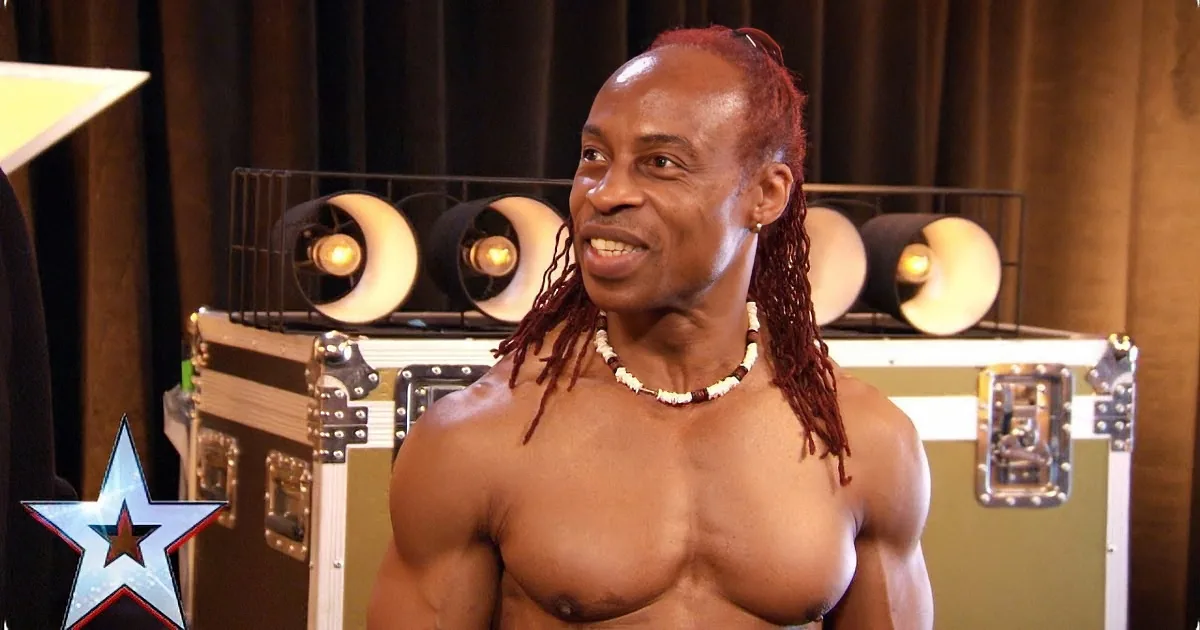A shirtless man with dreadlocks smiles in front of a backdrop with a star logo and stage lights.