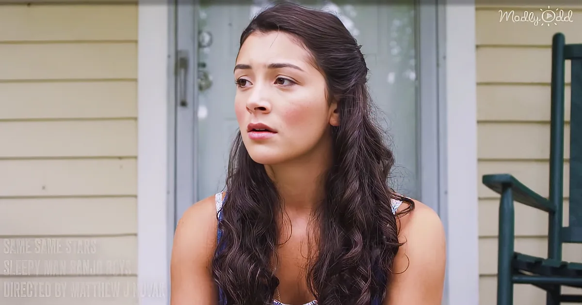 A woman with long dark hair and a surprised expression, wearing a sleeveless top, stands outside near a door and a green...
