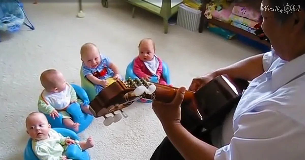 A person plays guitar while four babies in colorful seats watch attentively.