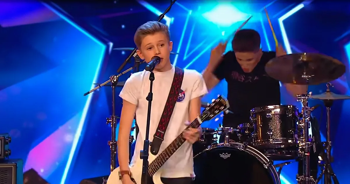 A young boy plays guitar on stage with a drummer behind him, under vibrant blue stage lights.