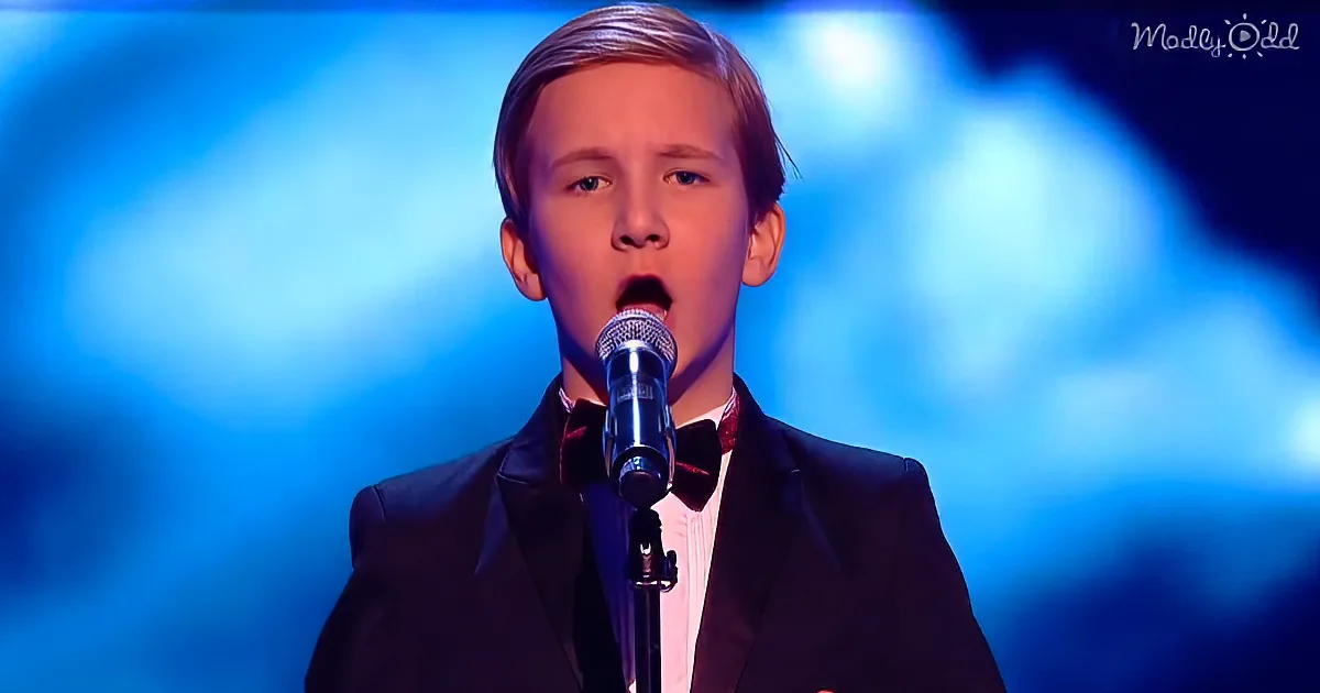 A young boy in a tuxedo sings into a microphone on stage with a blue background.