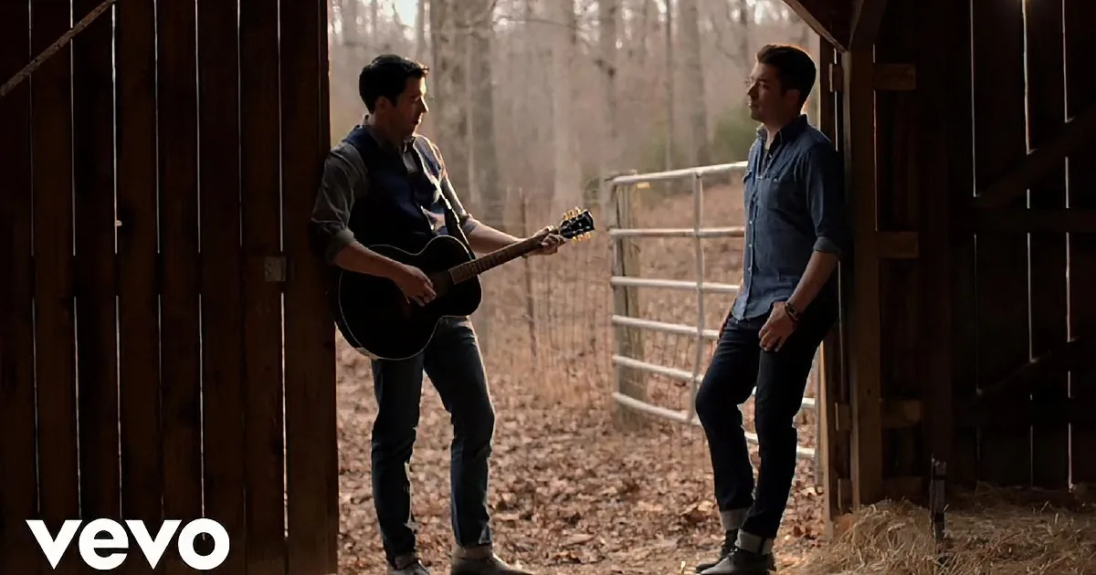 Two men in a barn, one playing guitar, the other leaning against the wall.