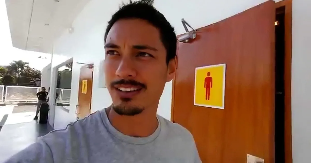 A man with a mustache and short hair smiles in front of a wooden door with a male restroom sign.