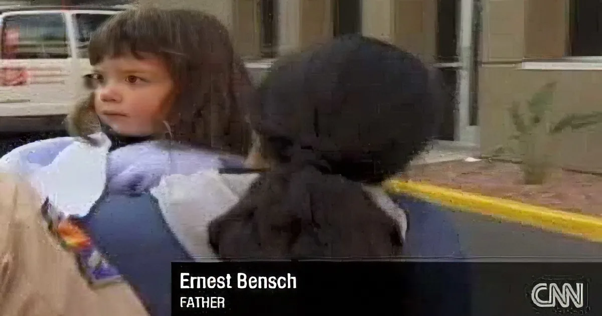 A young child with long hair is being held by a person in a dark coat, with a vehicle and building in the background.