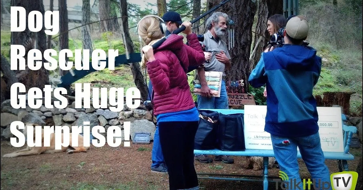 A dog rescuer receives a surprise, surrounded by filming equipment and a large check.