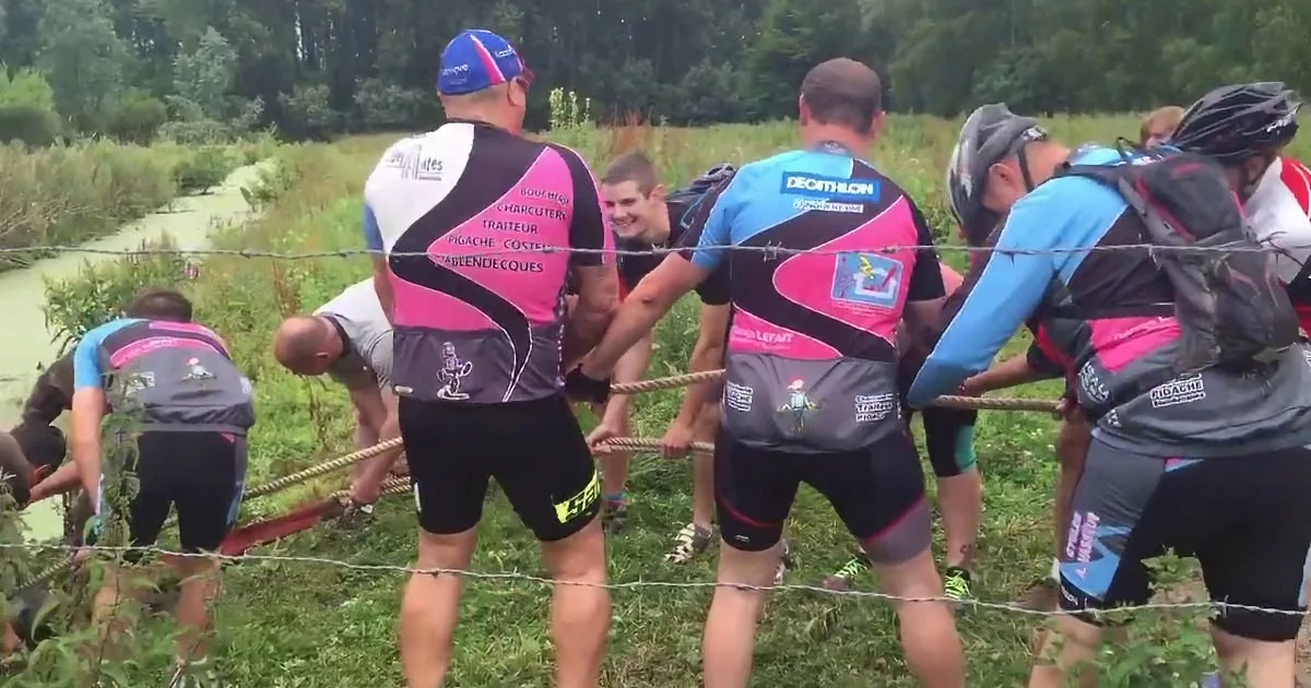 Athletes in pink and blue jerseys pull a rope over a barbed wire fence in a grassy field.