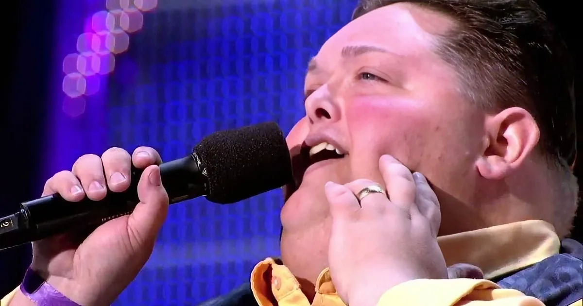 A man sings passionately into a microphone, wearing a ring and a yellow shirt, with a blurred stage background.