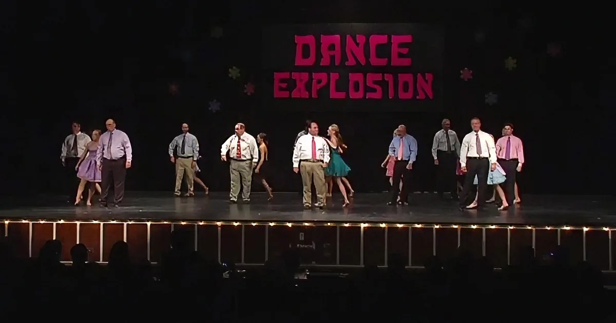 A group of people in formal attire dances on stage with "Dance Explosion" displayed behind them.