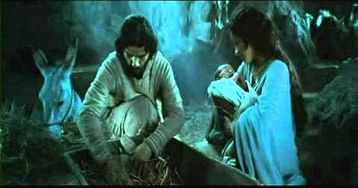 Mary holds baby Jesus while Joseph sits nearby in a dimly lit stable.