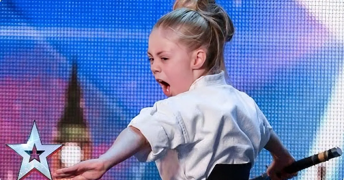 A young girl in a white shirt performs on stage, holding a microphone, with a colorful background featuring a star and a...