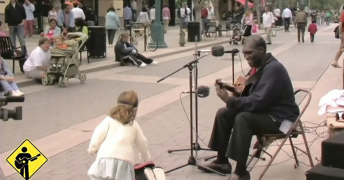 A man plays guitar on a sidewalk as a young girl approaches, surrounded by onlookers.