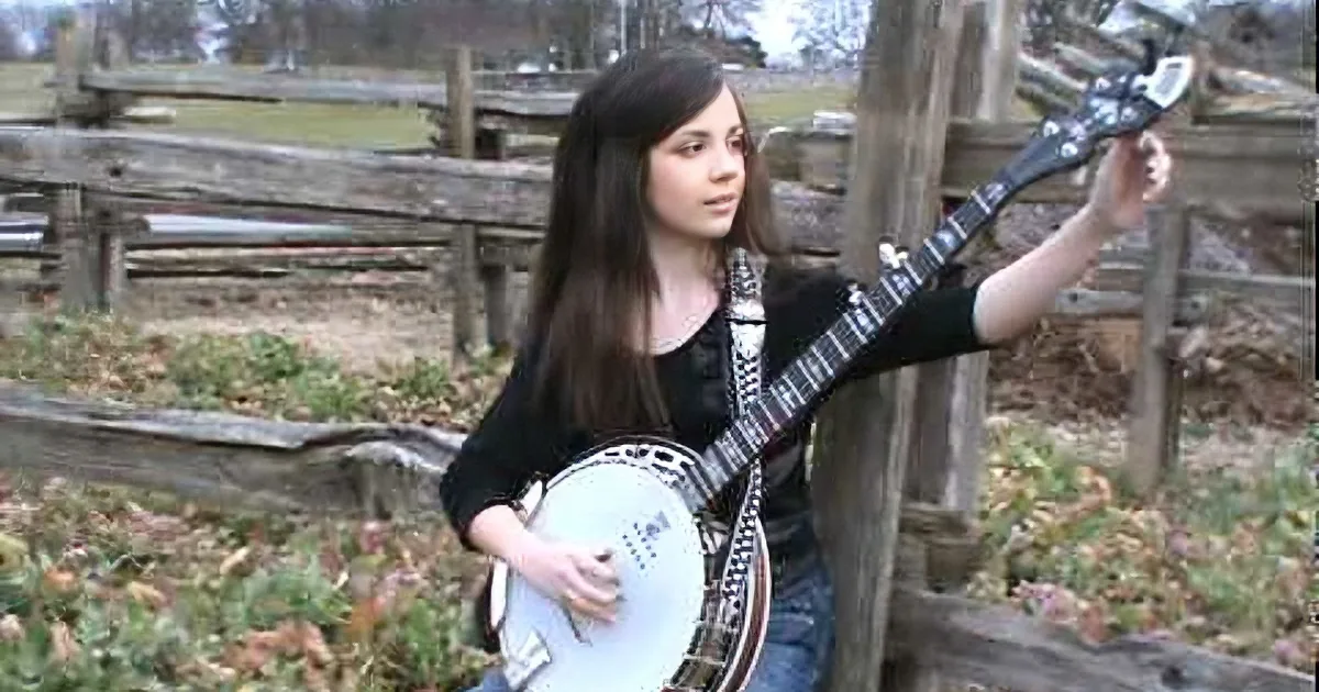 A girl with long dark hair plays a banjo in front of a rustic wooden fence.