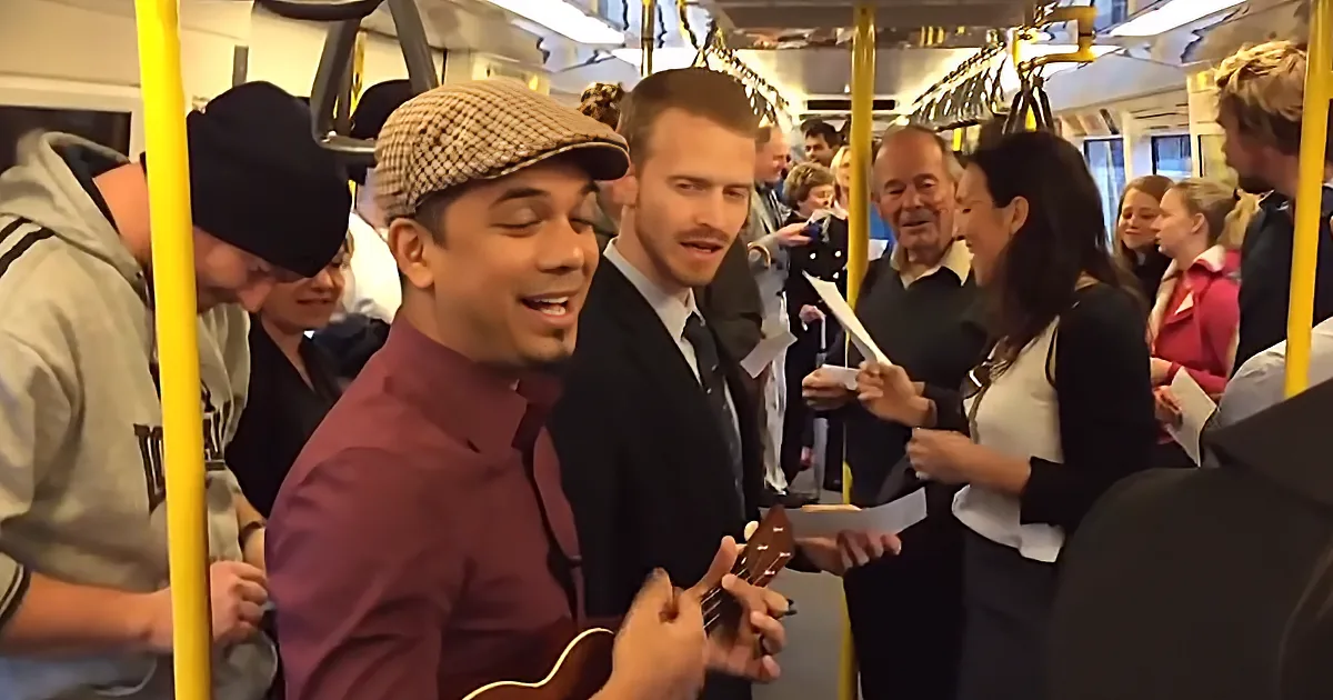 A man in a maroon shirt and hat plays guitar on a crowded bus, surrounded by passengers.