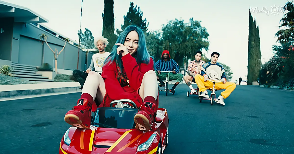 A person in a red outfit rides a small red car, surrounded by others on tricycles in a suburban setting.