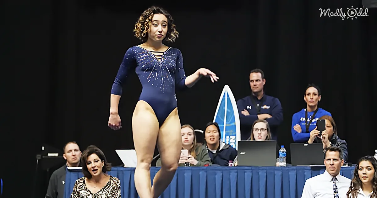 A gymnast in a blue leotard performs on a balance beam, with spectators and judges in the background.