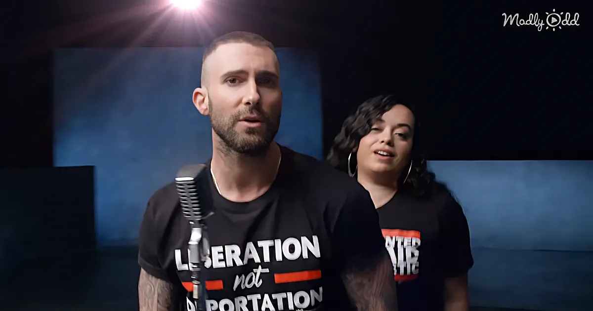 A man with a beard and tattoos wears a "Liberation not Deportation" shirt, standing in front of a microphone.