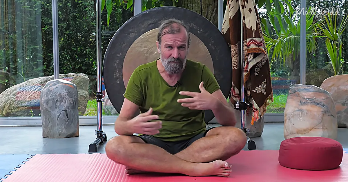 A man sits cross-legged on a mat, gesturing with his hands in front of a large gong and rocks.