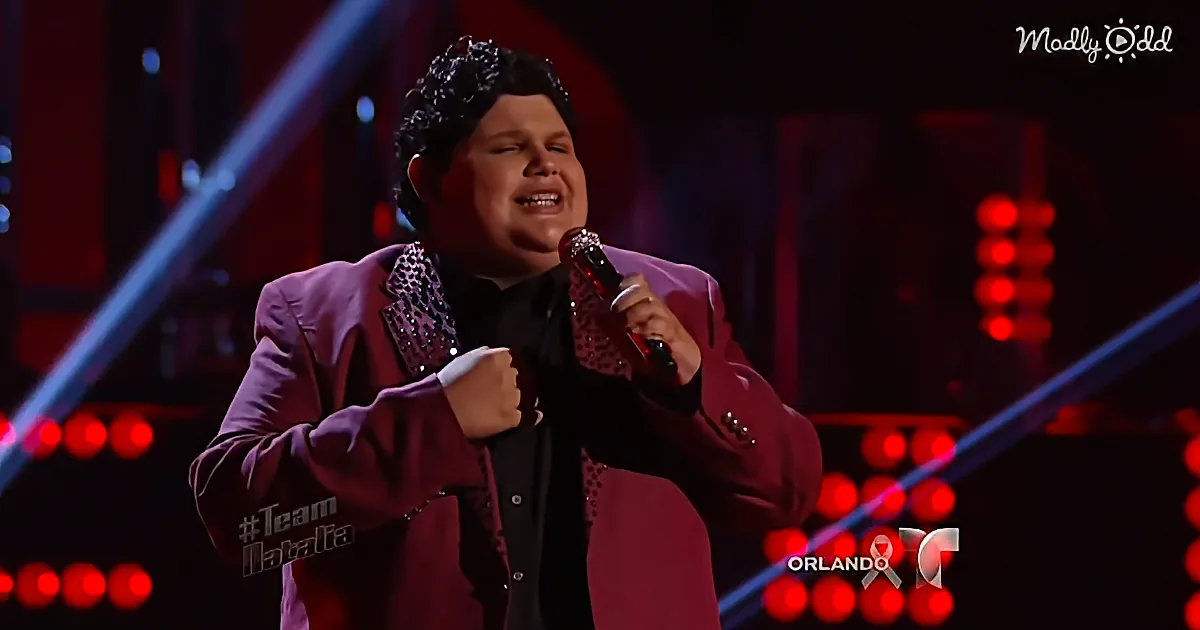 A person in a purple suit sings passionately on stage with red lights and a microphone.