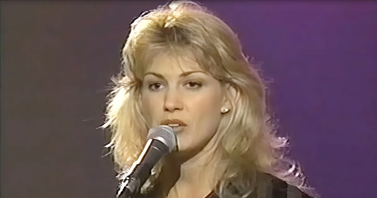 A woman with blonde hair singing into a microphone, wearing a black top and earrings.