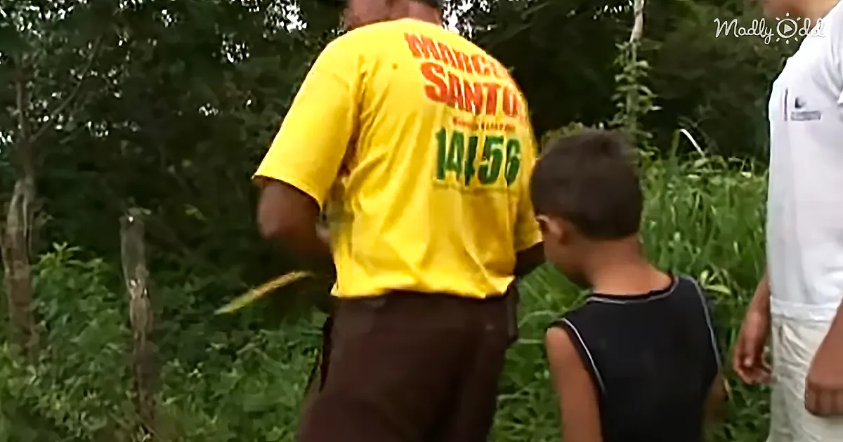 A man in a yellow shirt with "SANTO 1456" stands with a child and another person in a green forested area.