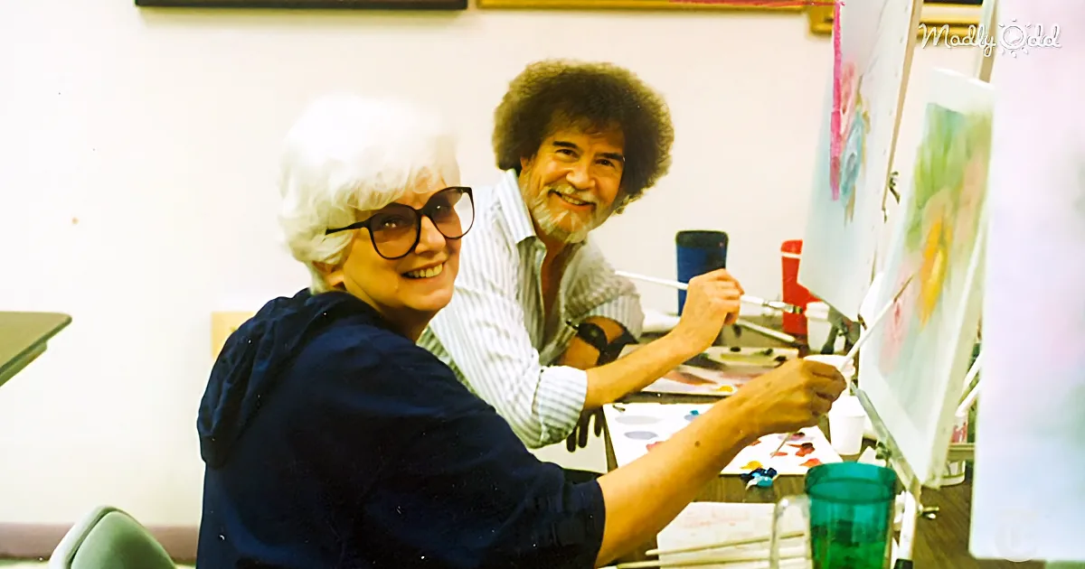 A woman in a blue shirt smiles while painting with Bob Ross, who is also smiling and holding a paintbrush.