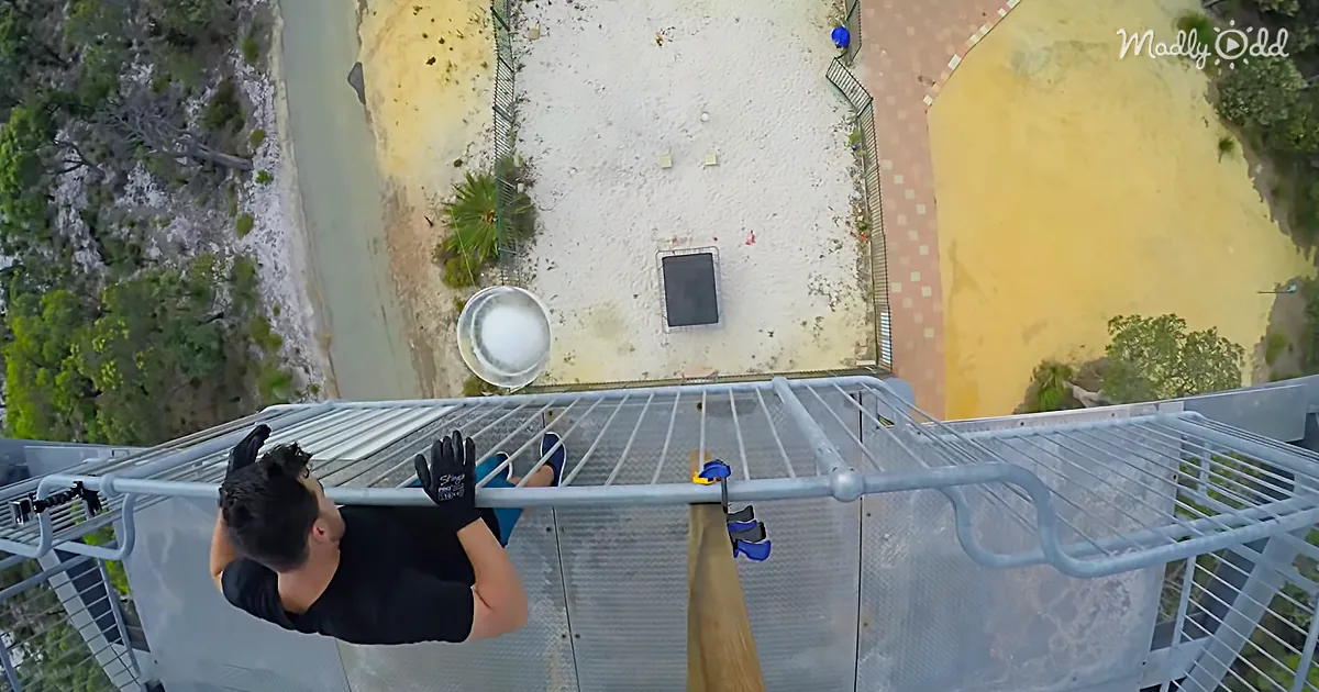 A person in black gloves stands on a metal platform, looking down at a bucket below, with a sandy area and trees in the...