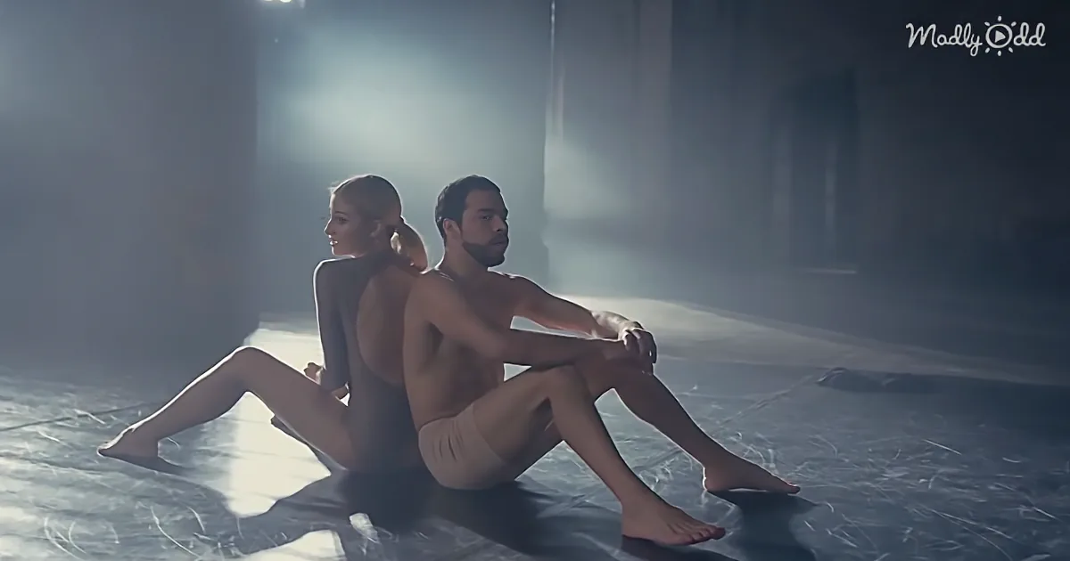 A man and woman sit back-to-back on a dark floor, illuminated by a spotlight, creating a dramatic and intimate scene.