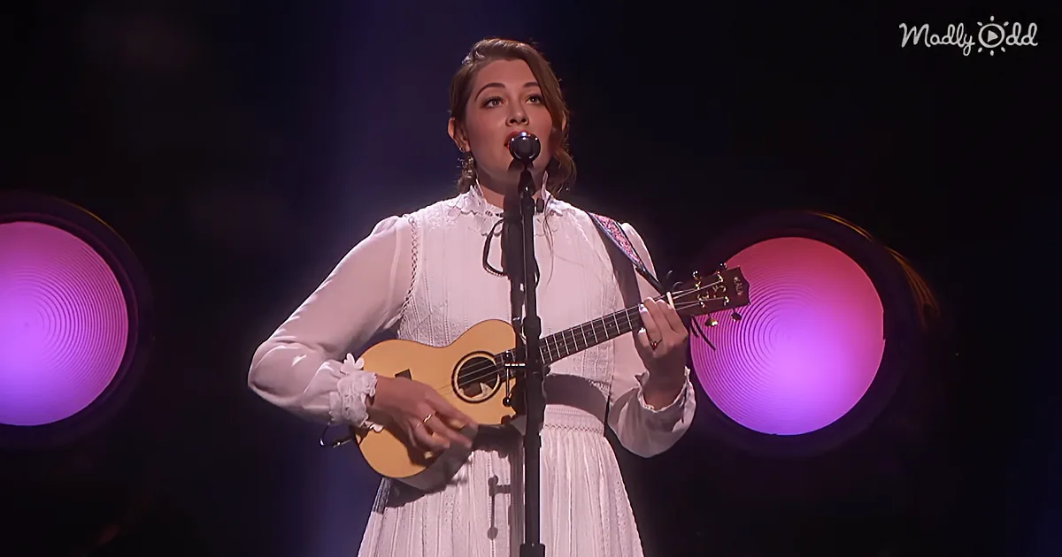 A woman in a white dress plays a ukulele and sings into a microphone on stage with purple lights.
