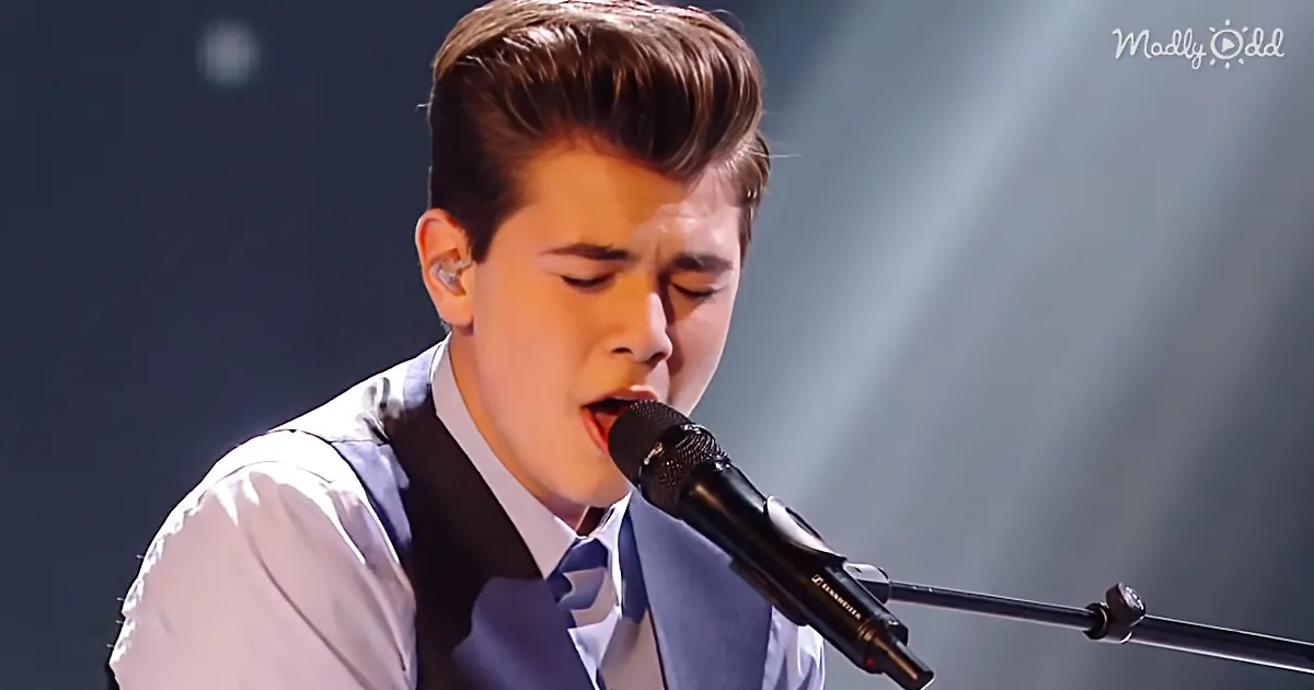 A young man in a suit sings into a microphone on stage under spotlight.