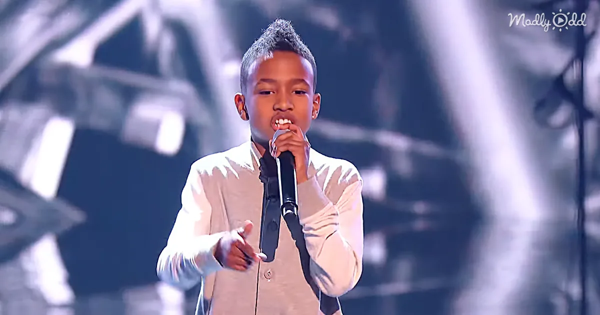 A young boy in a white shirt sings into a microphone on stage, gesturing with his hand.