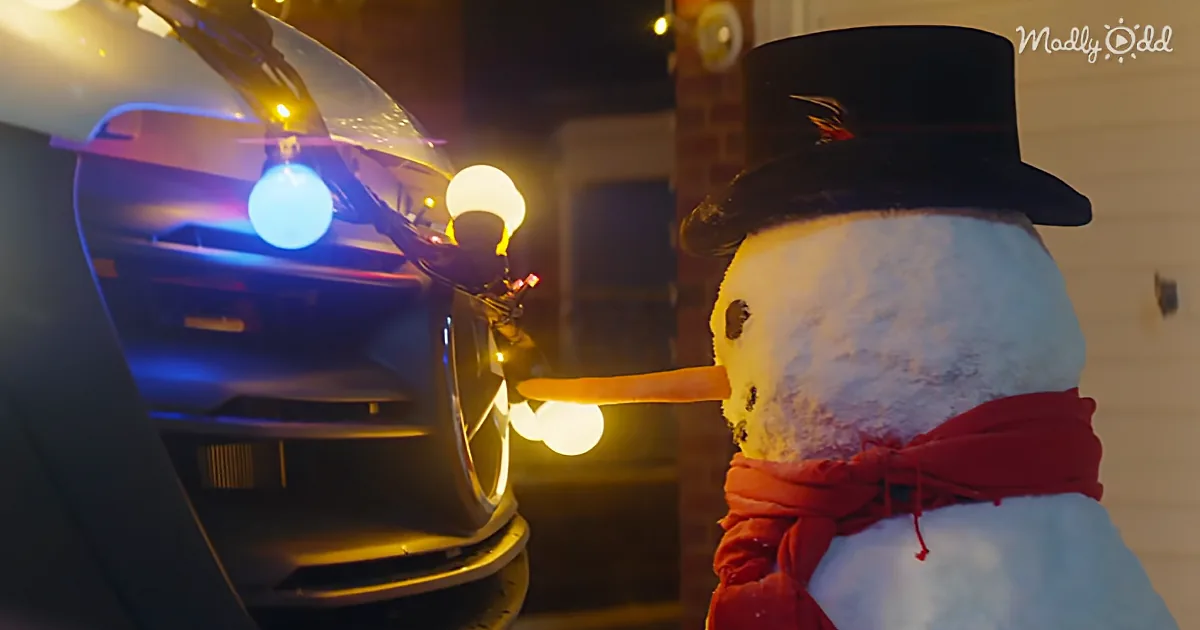 A snowman with a top hat and scarf touches a car's front bumper under a streetlight.