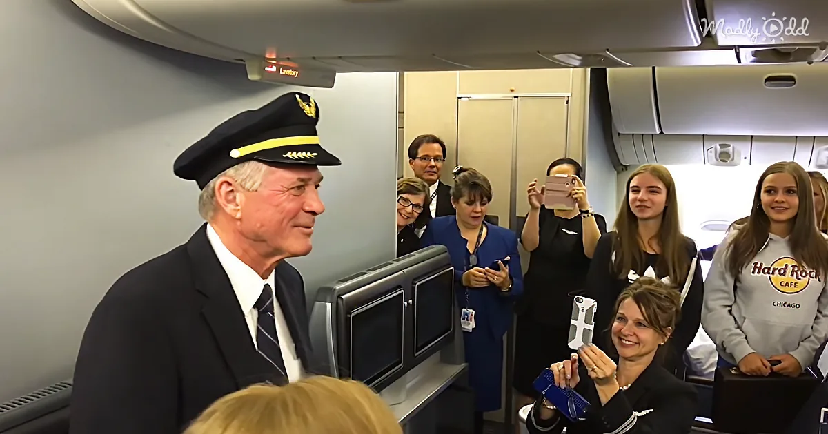 An older man in a pilot's uniform is smiling at a group of people taking photos inside an airplane.