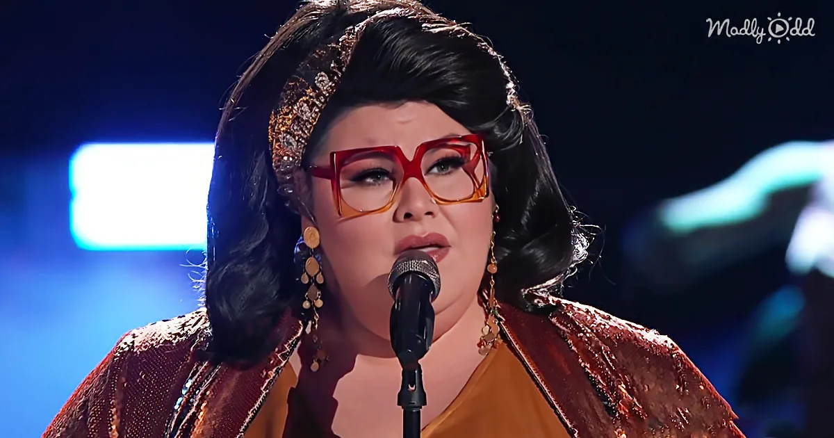 A woman with dark hair, red glasses, and a red outfit sings into a microphone on stage.