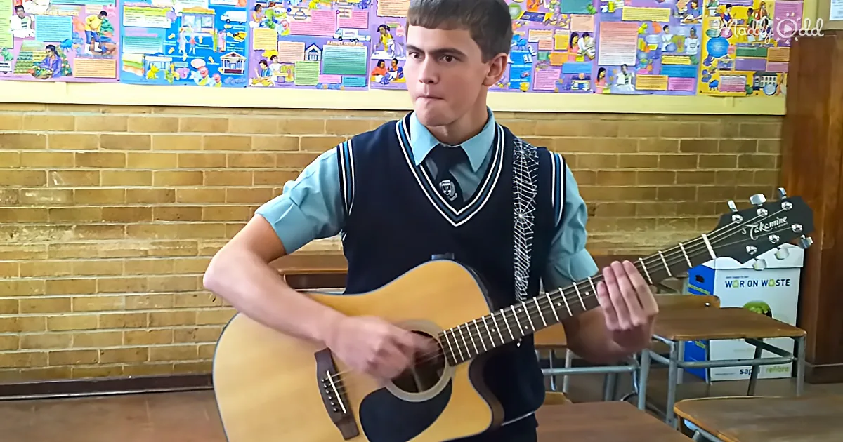 A young man in a school uniform plays an acoustic guitar in a classroom with colorful posters on the wall.