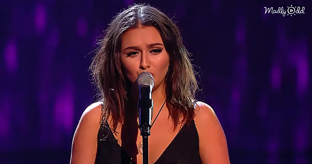 A woman with long hair and a nose ring sings into a microphone on stage with a purple background.