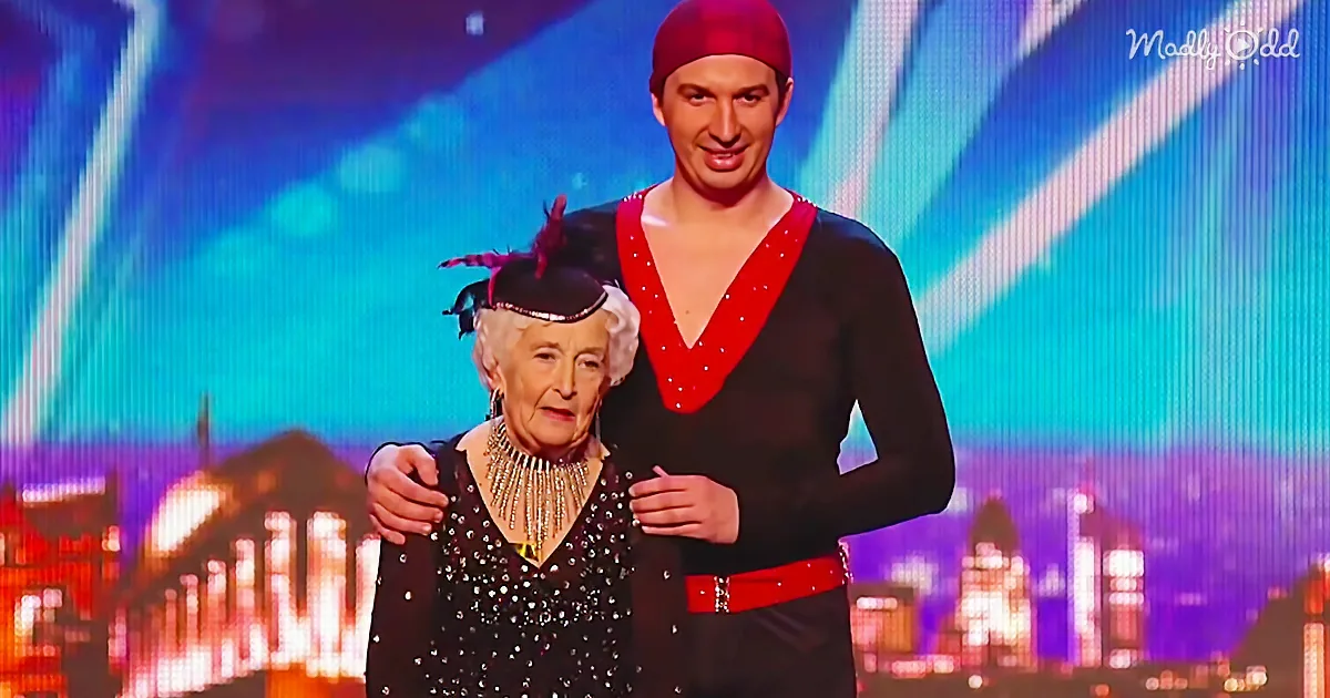 An elderly woman in a black dress with a feathered hat stands with a man in a red and black outfit on a stage with a...