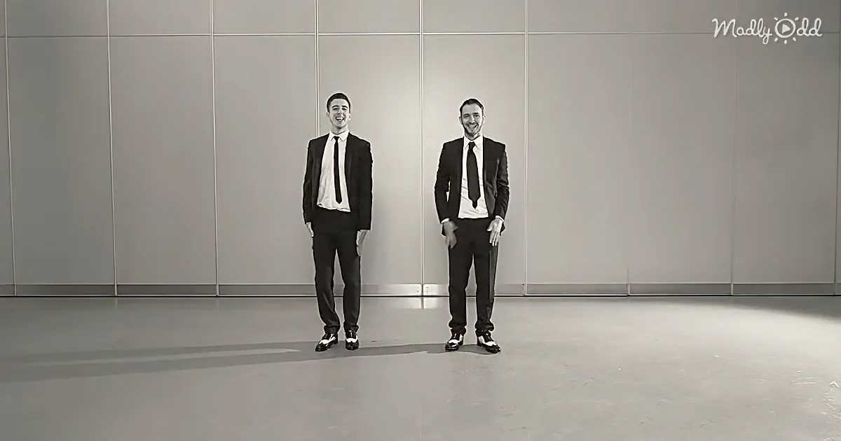 Two men in suits smile, standing side by side against a plain wall.