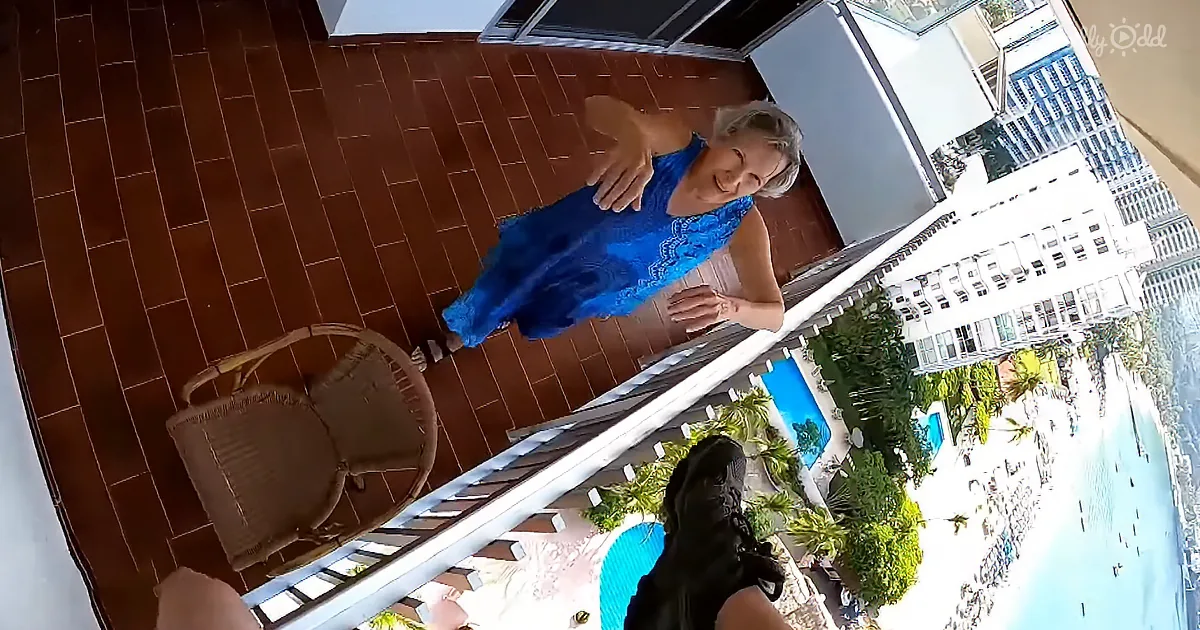 A woman in a blue dress stands on a balcony, smiling, with a wicker chair and a view of a pool and buildings below.