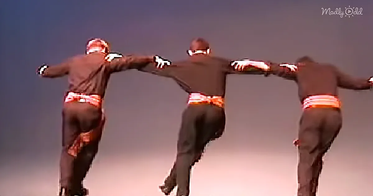 Three dancers in brown outfits with red belts perform synchronized moves against a gradient sky backdrop.