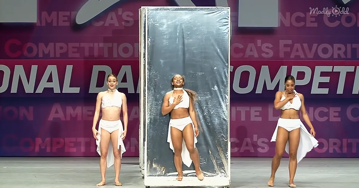 A woman in a white outfit stands on a stage, posing with a large, wrapped object behind her.
