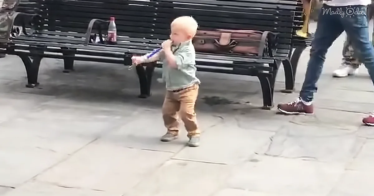 A toddler in a green shirt and brown pants stands on a stone pavement, holding a blue object near their mouth.