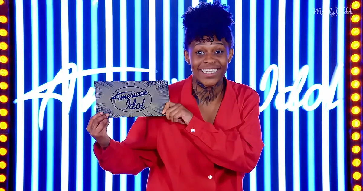 A person in a red outfit holds an "American Idol" sign, smiling, against a blue and white striped background.