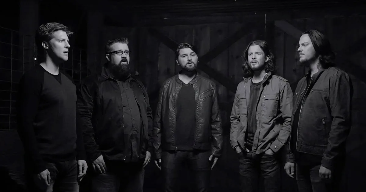 Five men in dark clothing stand together in a dimly lit, industrial setting.