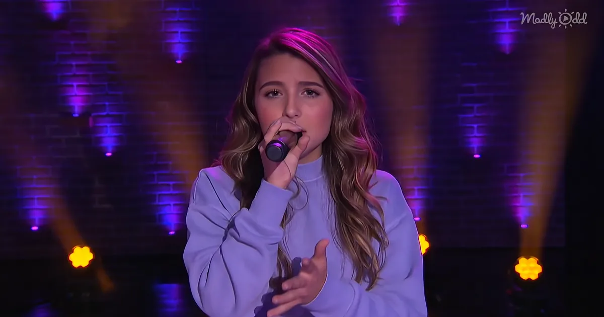 A person in a light blue outfit sings into a microphone, with a brick wall and colorful lights in the background.