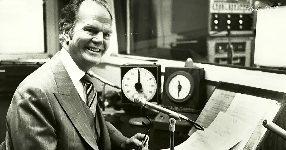 A man in a suit smiles while holding a pair of glasses, seated in front of a microphone and clock.